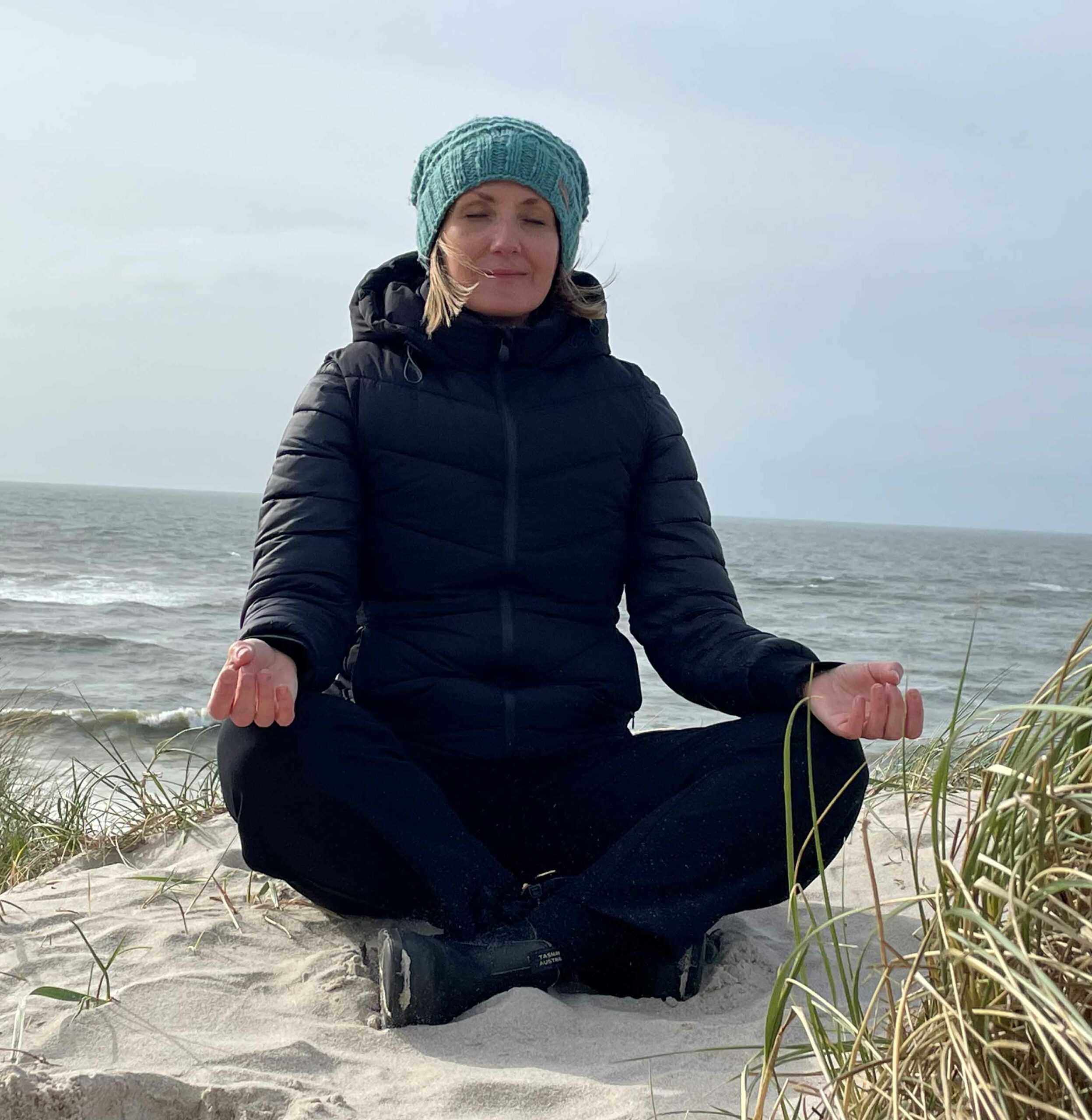 Frau sitzt im Schneidersitz auf einer Düne, im Hintergrund das Meer.